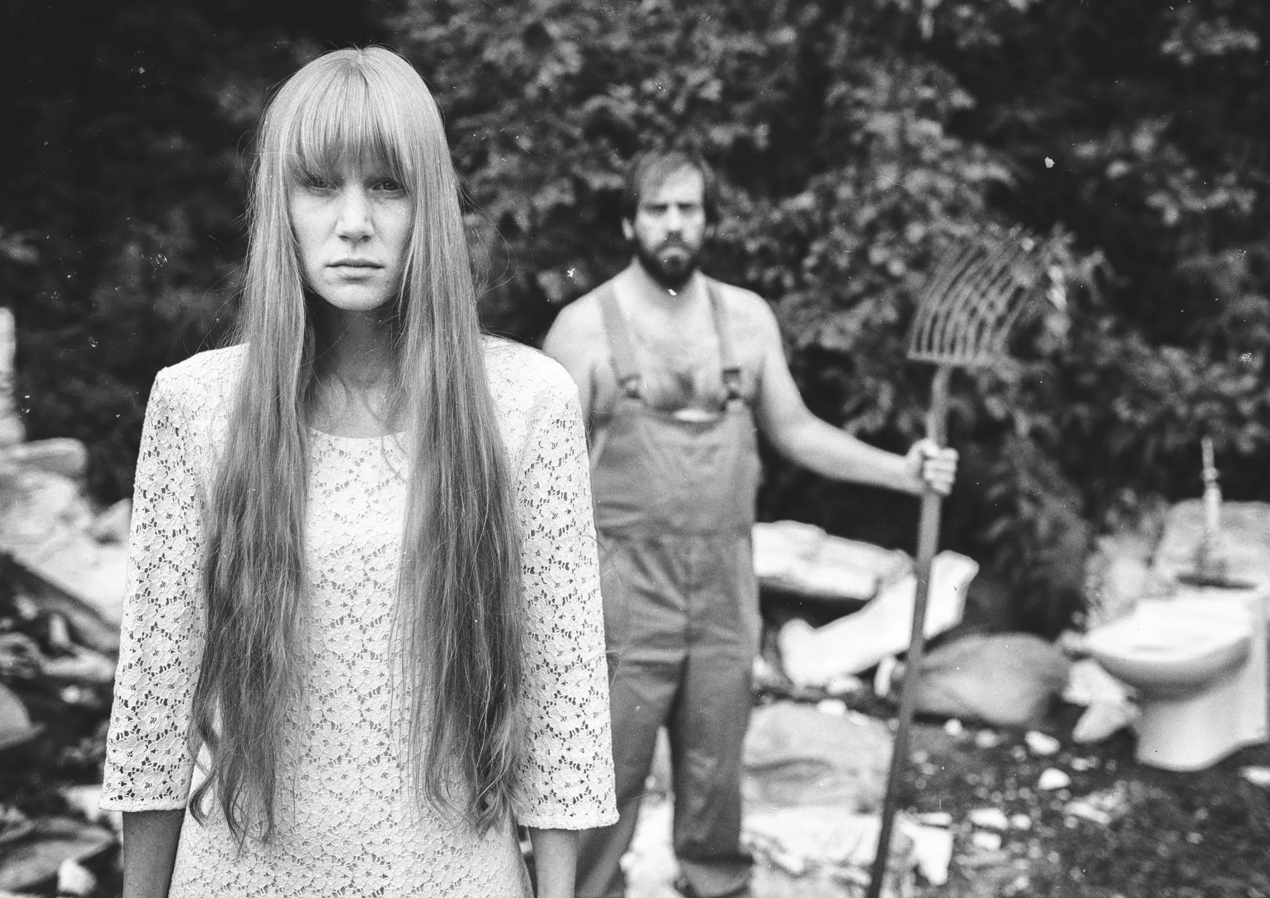 Black and white editorial shot of a woman with a man holding a pitchfork in the forest background.